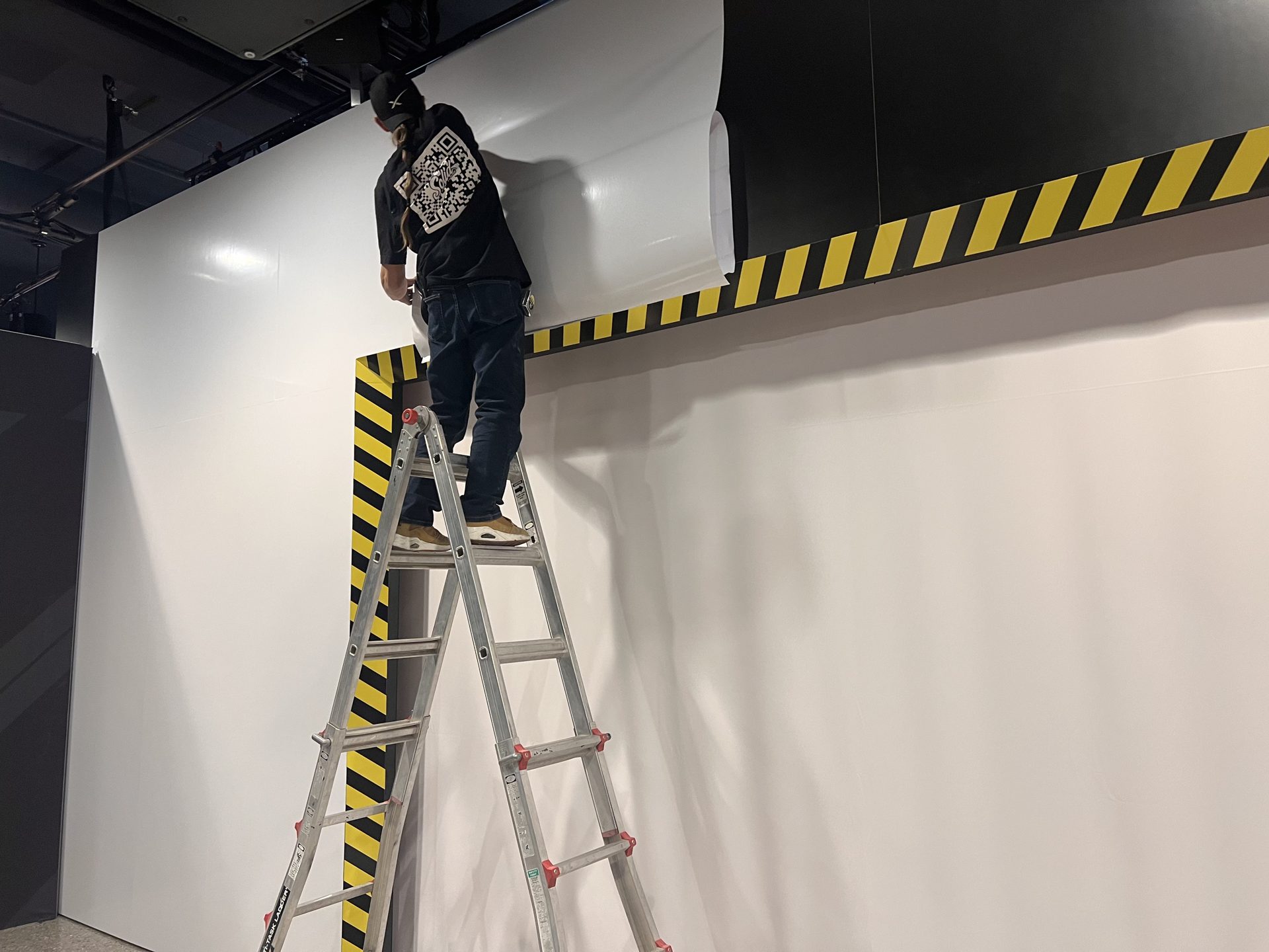 Seamless white wall wrap — ladder install above hazard-striped scenic deck